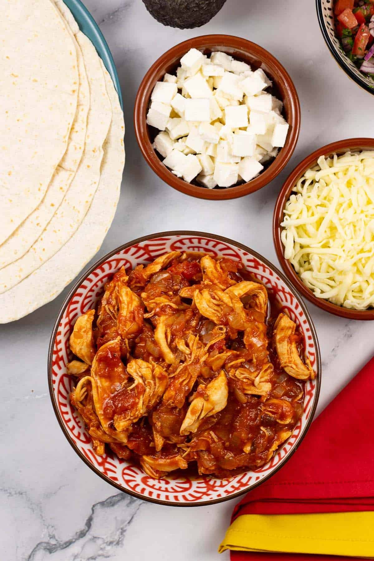 A bowl of chicken tinga filling with tortilla wraps and dishes of feta and mozzarella on the side.