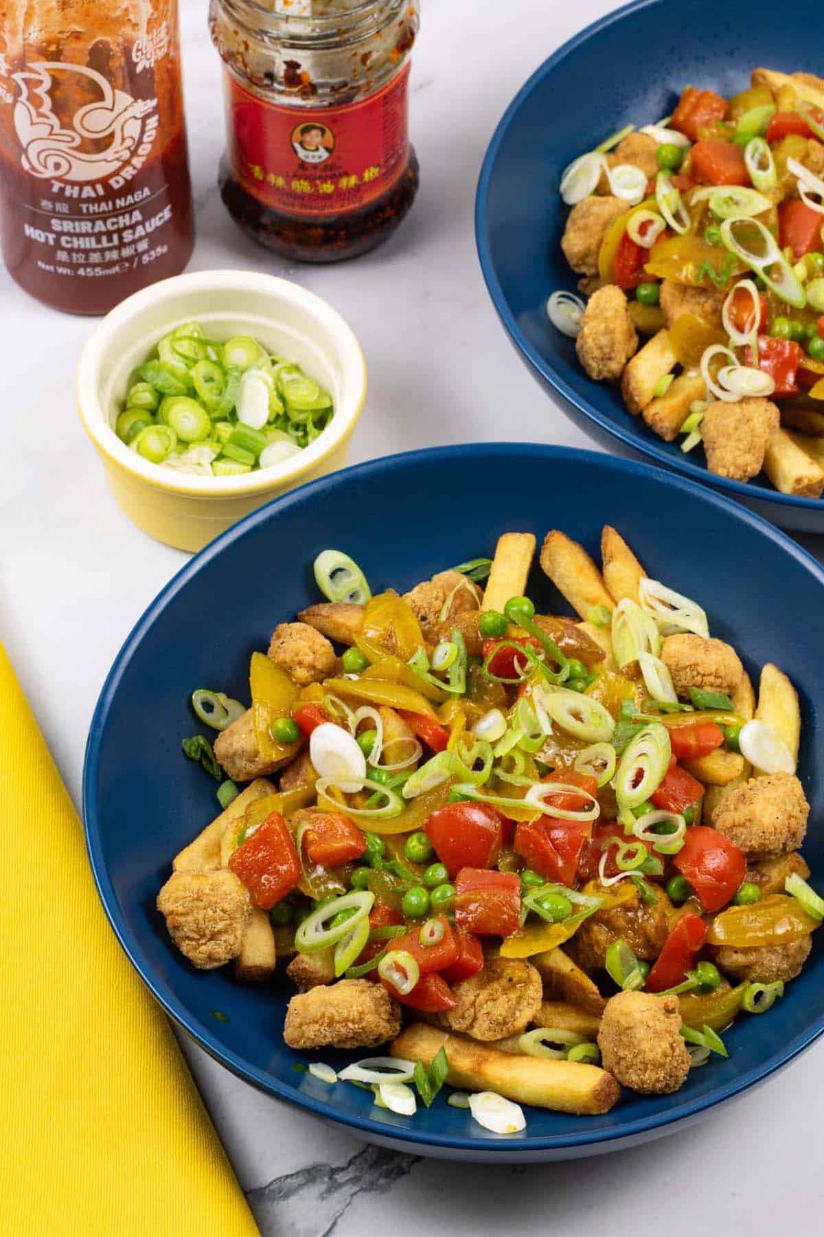 2 portions of crispy chicken curry loaded fries in dark blue bowls with a small dish of sliced spring onions and a yellow napkin on the side.