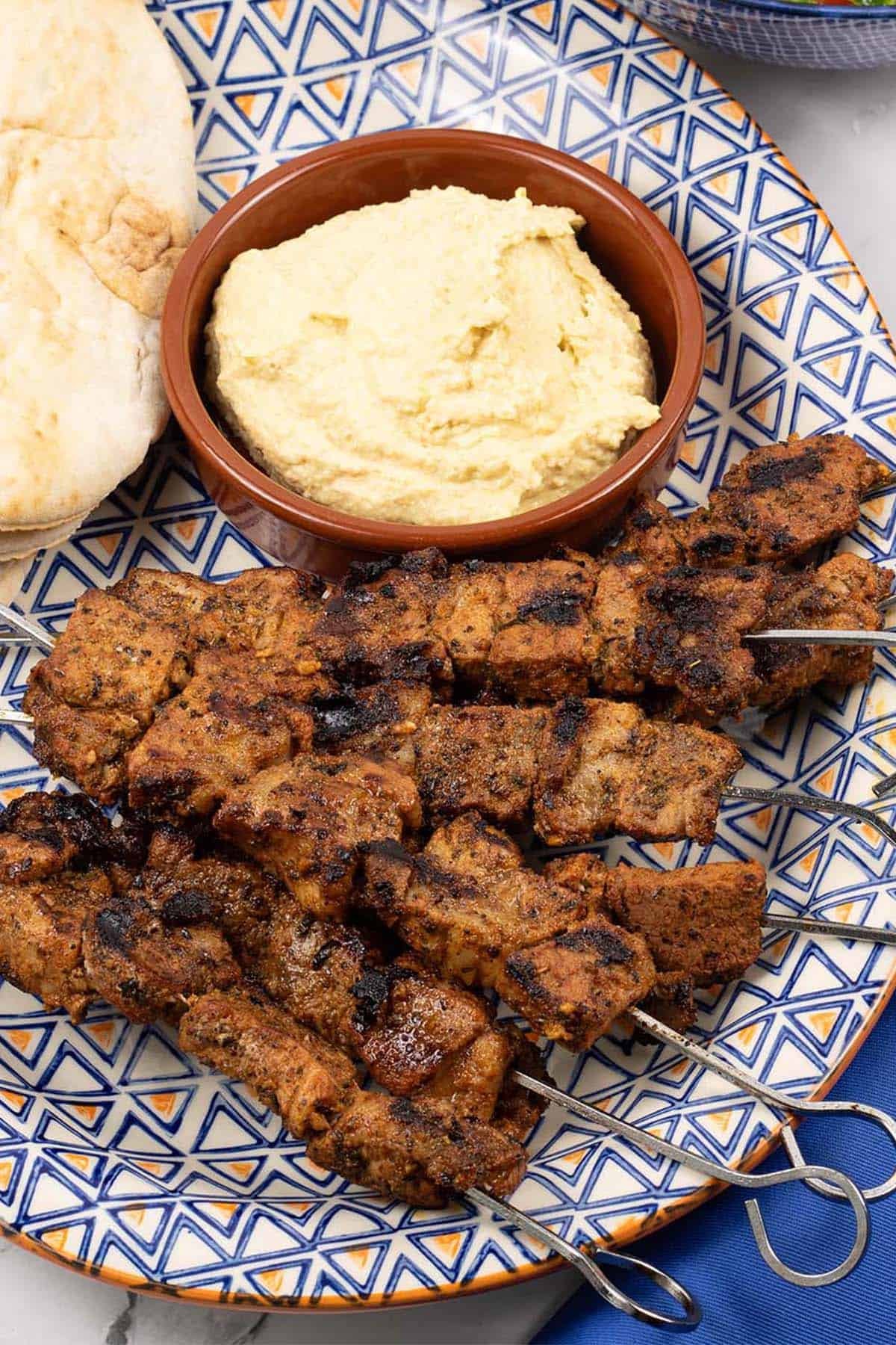 Easy greek pork skewers on an orange blue and white patterned platter with pitta breads and houmous.