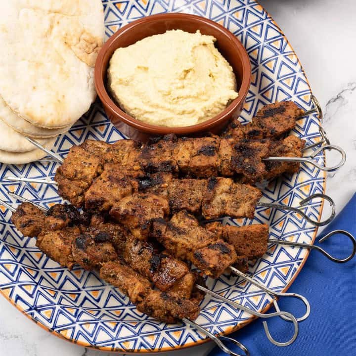 Easy greek pork skewers on an orange blue and white patterned platter with pitta breads and houmous.