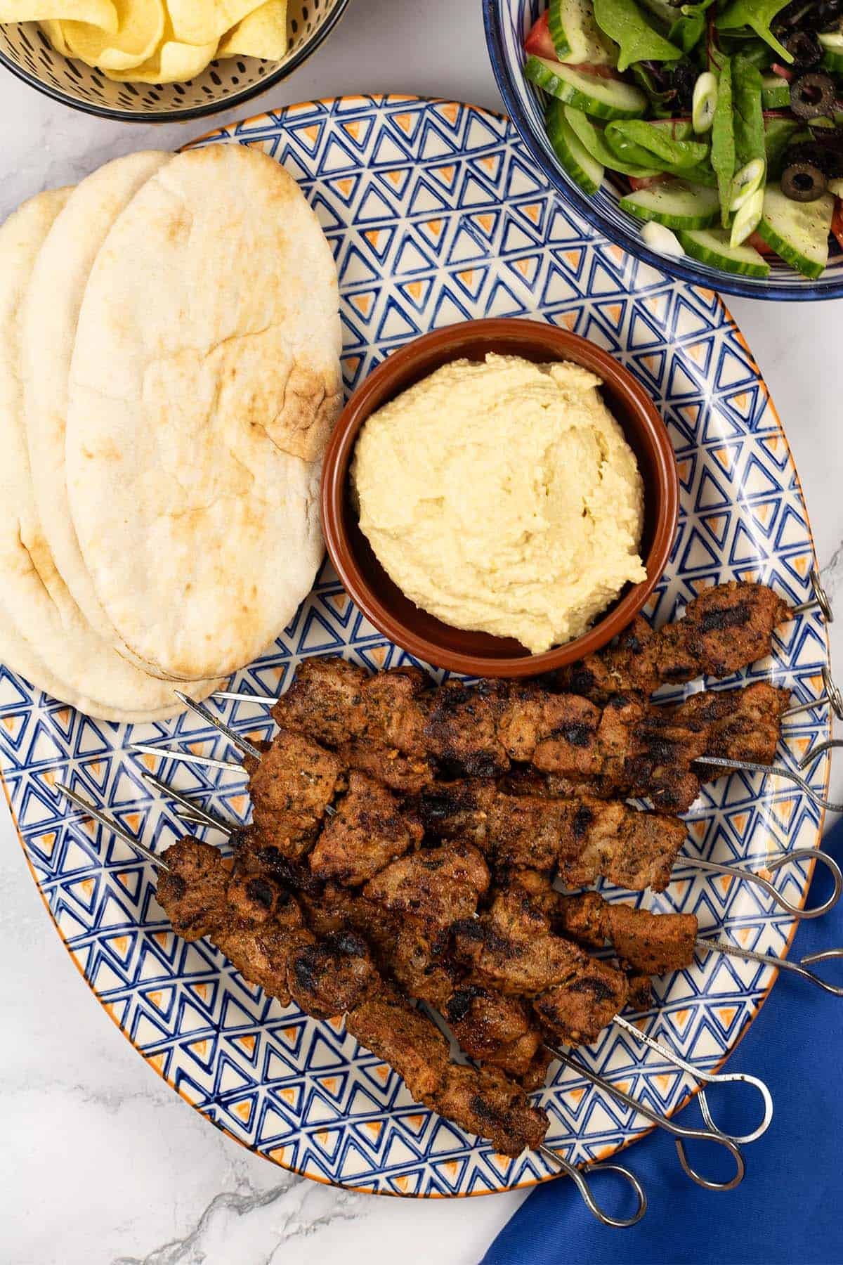 Easy greek pork skewers on an orange blue and white patterned platter with pitta breads and houmous.