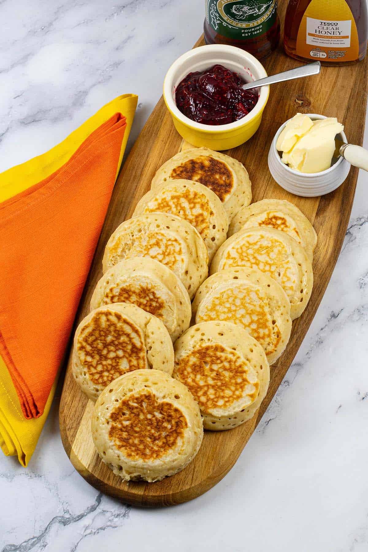 Homemade crumpets on a board with dishes of jam and butter, and bottles of syrup and honey, with an orange and yellow napkins on the side.