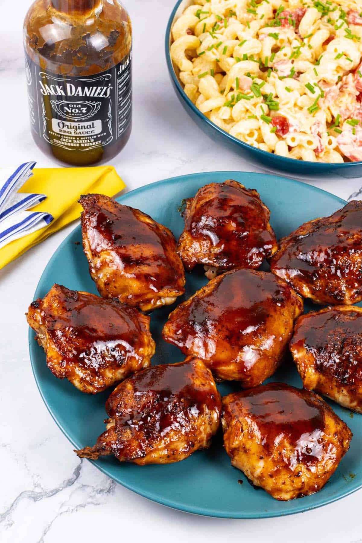 Juicy BBQ chicken thighs on a teal plate with a bottle of barbecue sauce and bowl of pasta salad on the side.