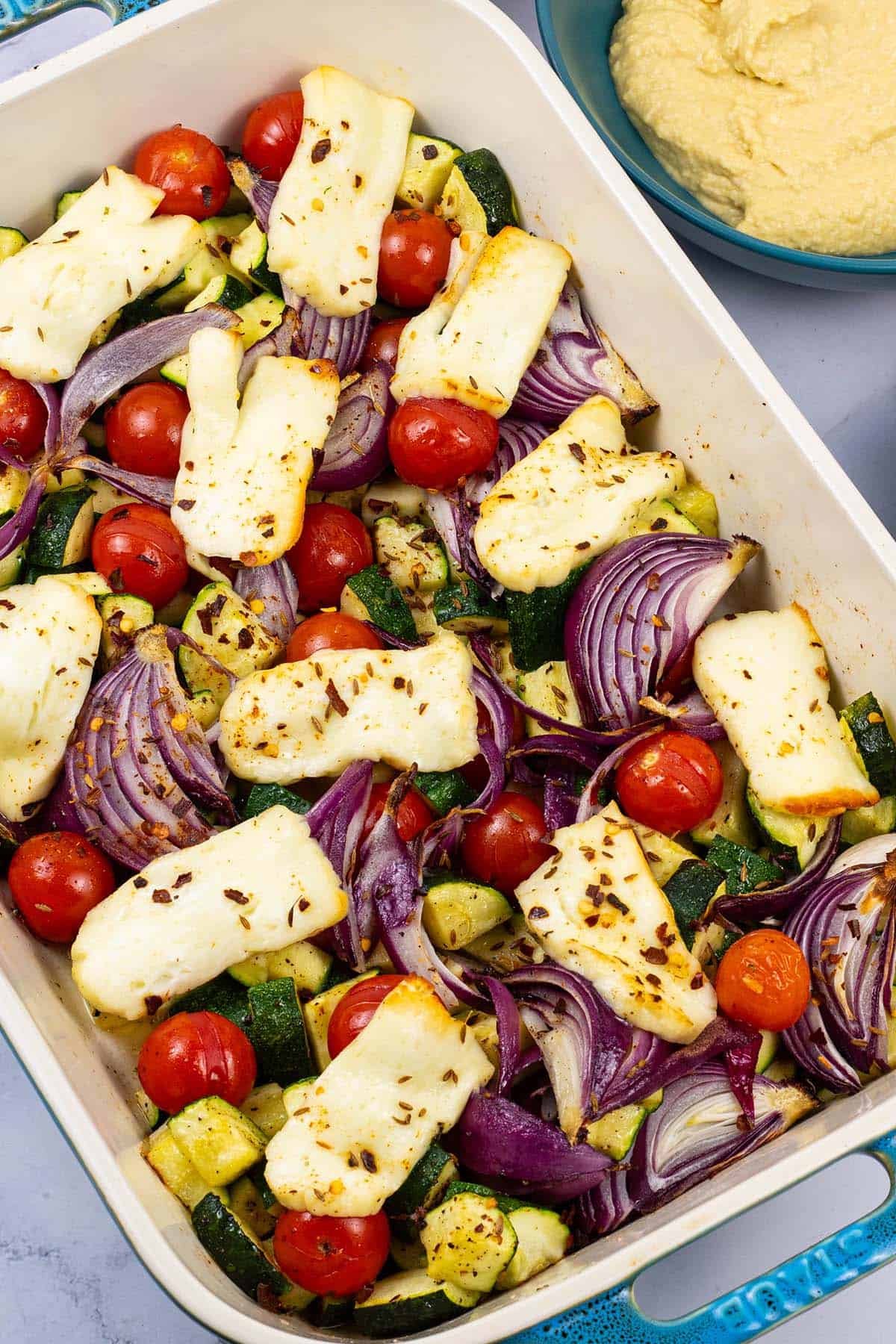 Halloumi and mediterranean veg tray bake in a large rectangle Staub dish, with houmous on the side.
