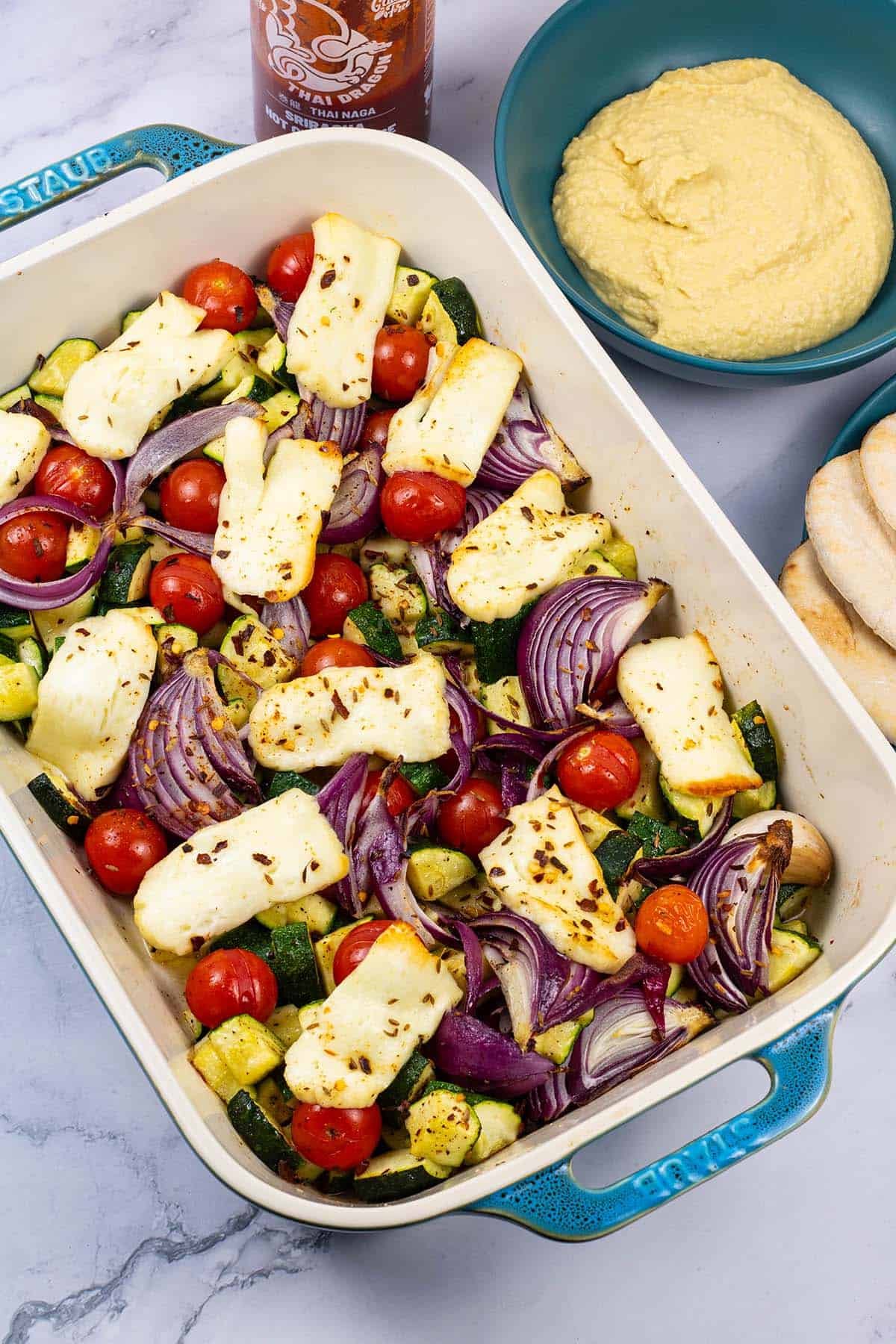 Halloumi and mediterranean veg tray bake in a large rectangle Staub dish, with pitta breads, houmous and sriracha on the side.