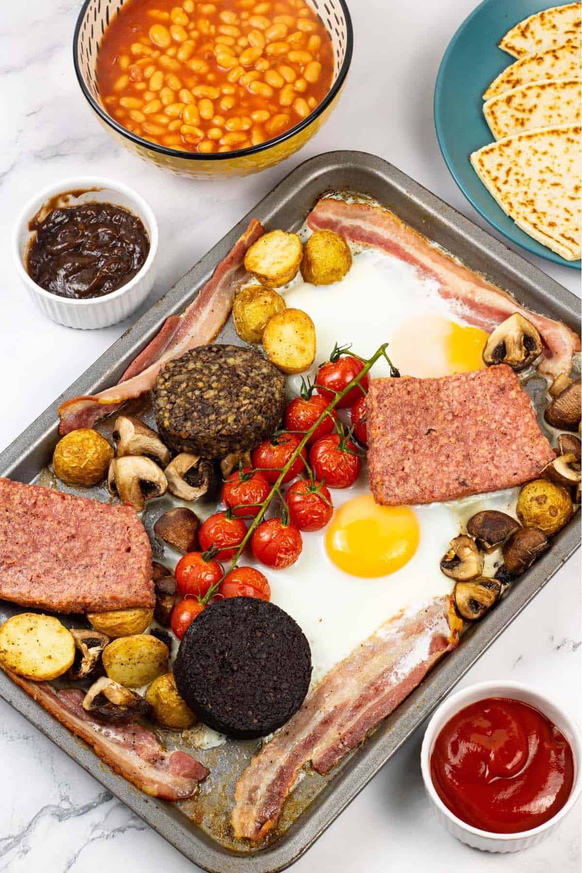 Scottish breakfast traybake on a baking tray with a bowl of baked beans, plate of tattie scones and small dishes of brown and tomato sauces on the side.
