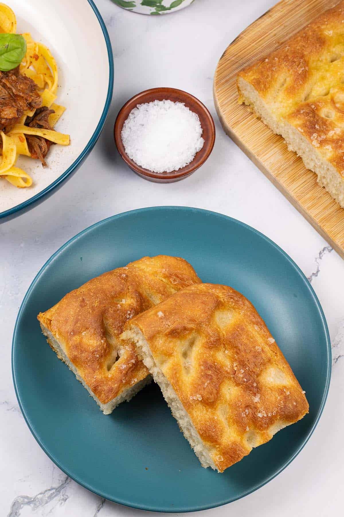 Two slices of authentic focaccia on a teal side plate with the rest on a board and a bowl of slow cooker beef ragu in the background.