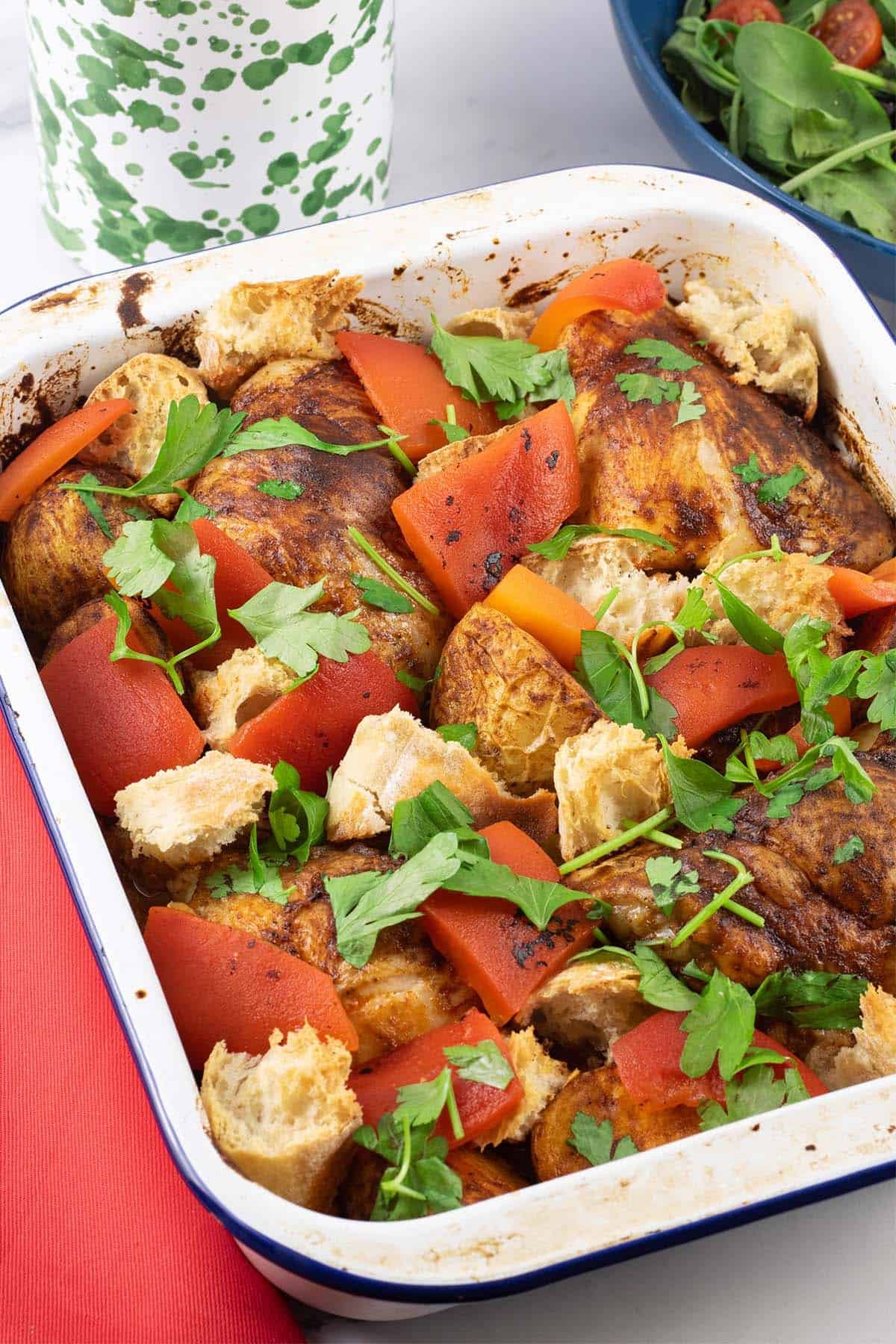 Chicken thighs with romesco traybake in a white enamel roasting tin with a bottle of oil and bowl of salad in the background.