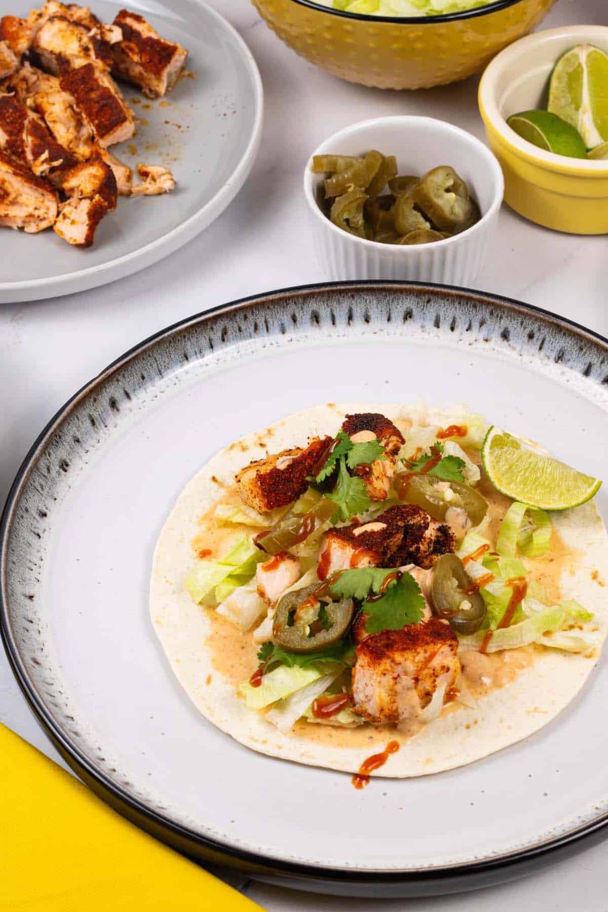 Grilled salmon tacos with chipotle lime yogurt on a white and black dinner plate with dishes of salmon, jalapeños, lettuce and lime wedges in the background.