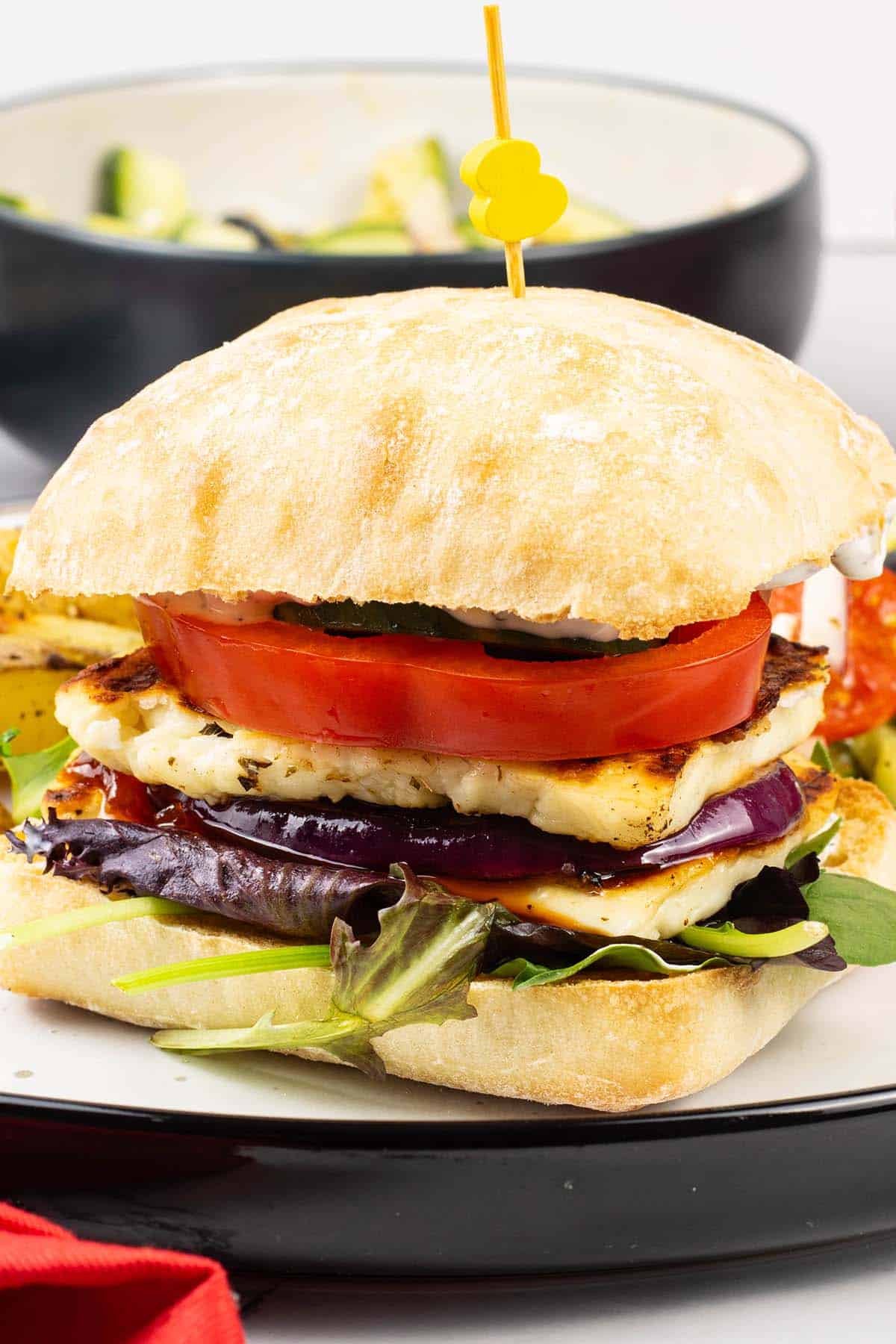 Halloumi burger on a white and black dinner plate with chunky oven chips and greek salad.