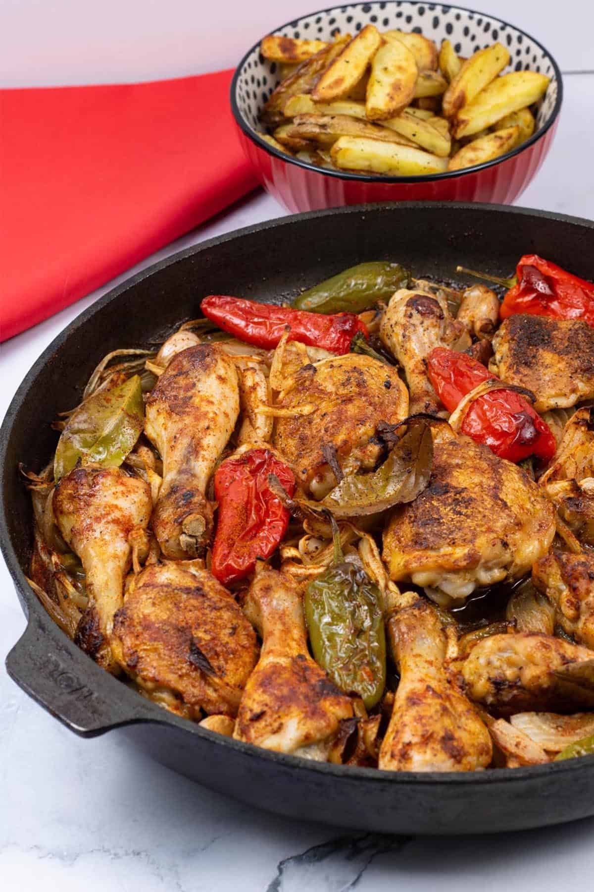 Paprika roast chicken in a large frying pan with a bowl of chunky oven chips and red napkin in the background.