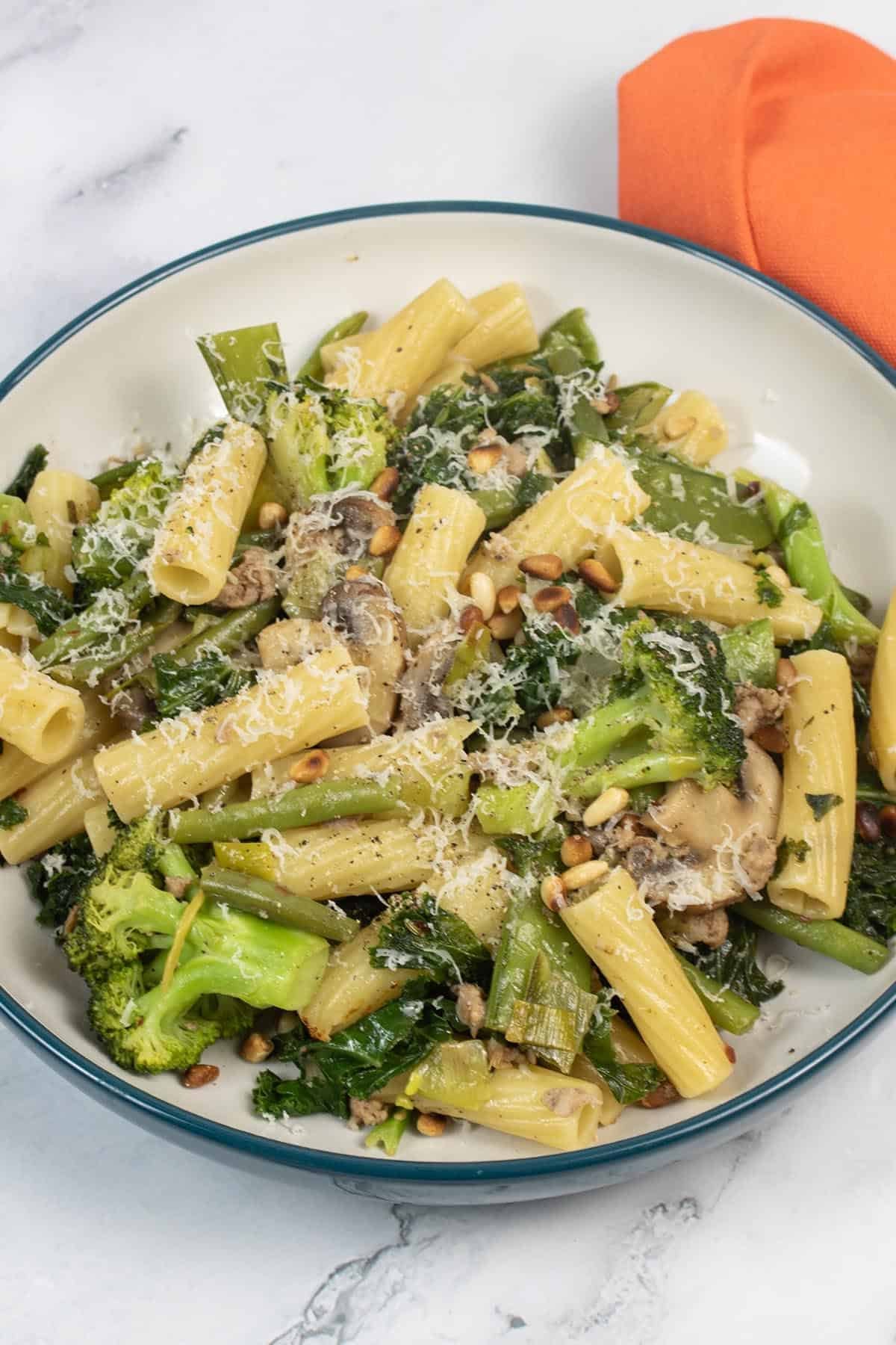 Sausage, vegetable and pine nut pasta in a bowl with an orange napkin on the side.