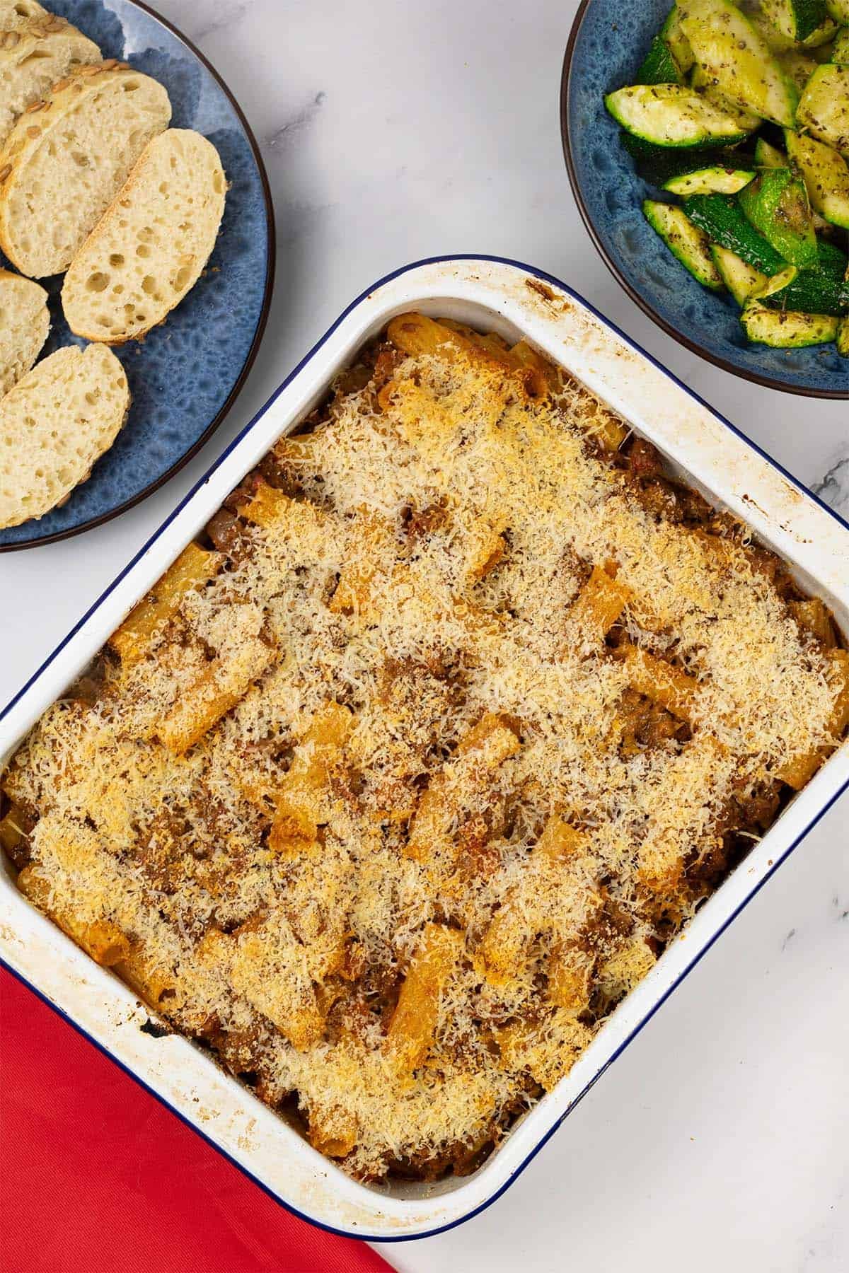 Beef and tomato pasta bake in a blue rimmed white enamel roasting tin with a bowl of air fryer courgettes and plate of sliced crusty bread in the background.