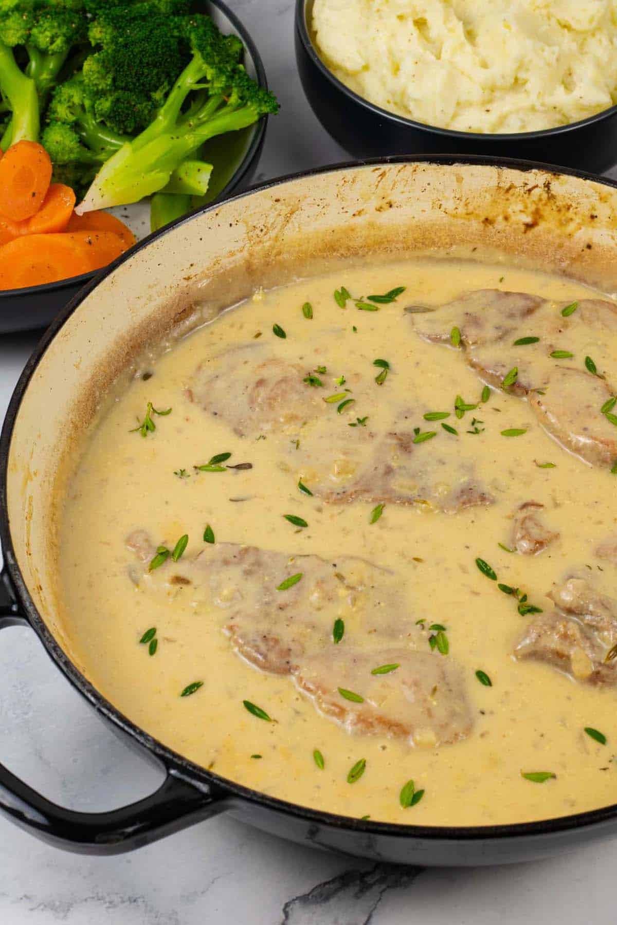 Braised pork steaks with mustard cream sauce in a shallow casserole with bowls of mashed potatoes, broccoli and carrots in the background.