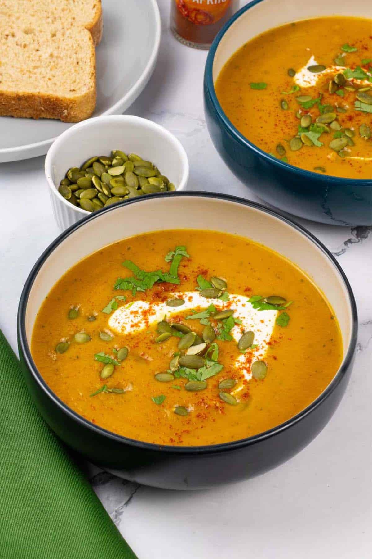 Two bowls of carrot and coriander soup with a small dish of toasted pumpkin seeds and plate of bread in the background.