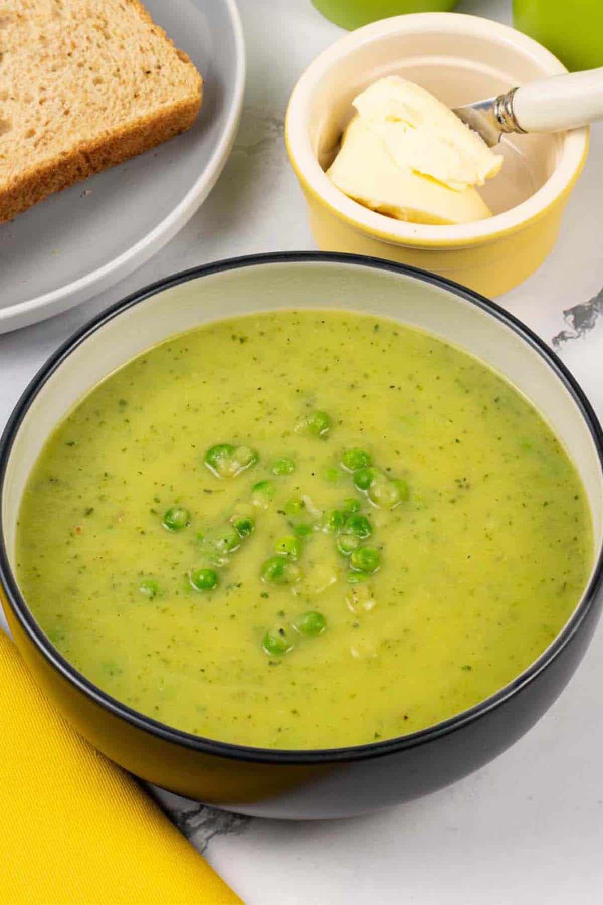 Courgette, potato and leek soup in a black and cream bowl with a plate of sliced bread and dish of butter in the background.