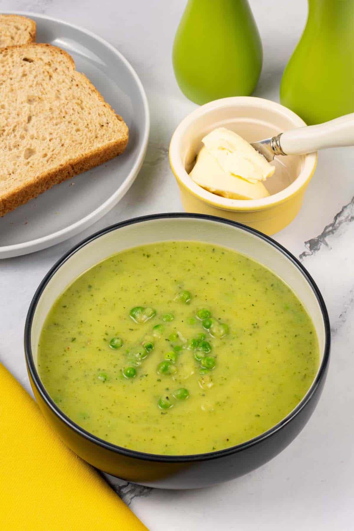 Courgette, potato and leek soup in a black and cream bowl with a plate of sliced bread, dish of butter and salt and pepper mills in the background.