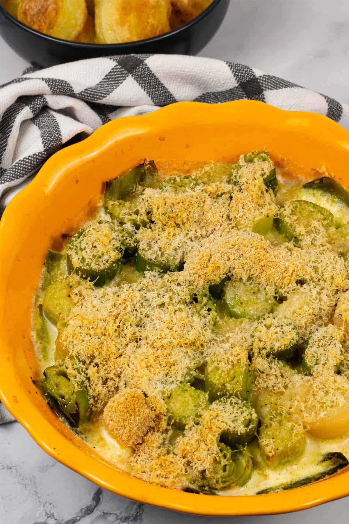 Leek gratin in a round yellow ovenproof dish with a bowl of roast potatoes in the background.