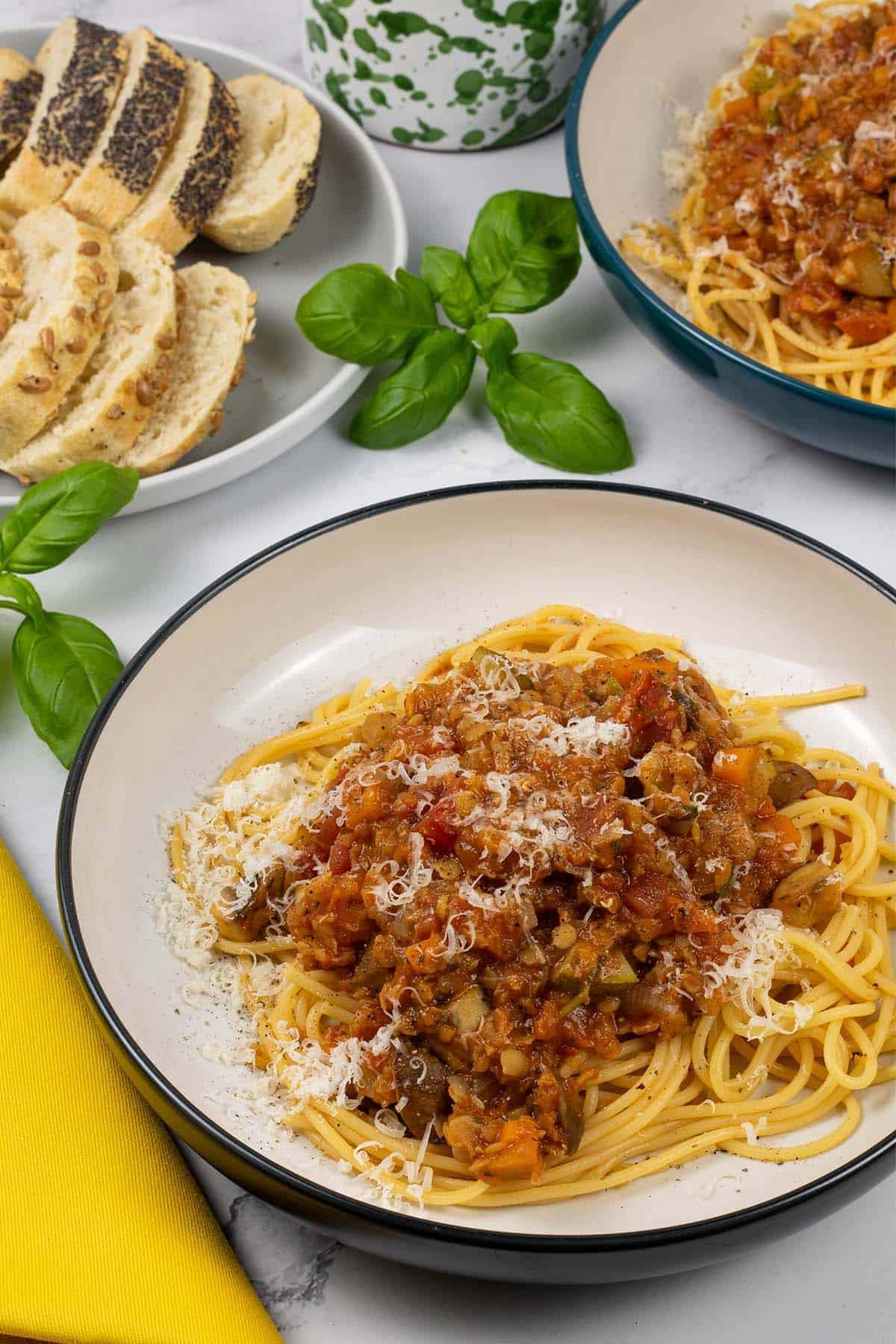 Two bowls of lentil and vegetable bolognese with spaghetti and a plate of sliced bread in the background.