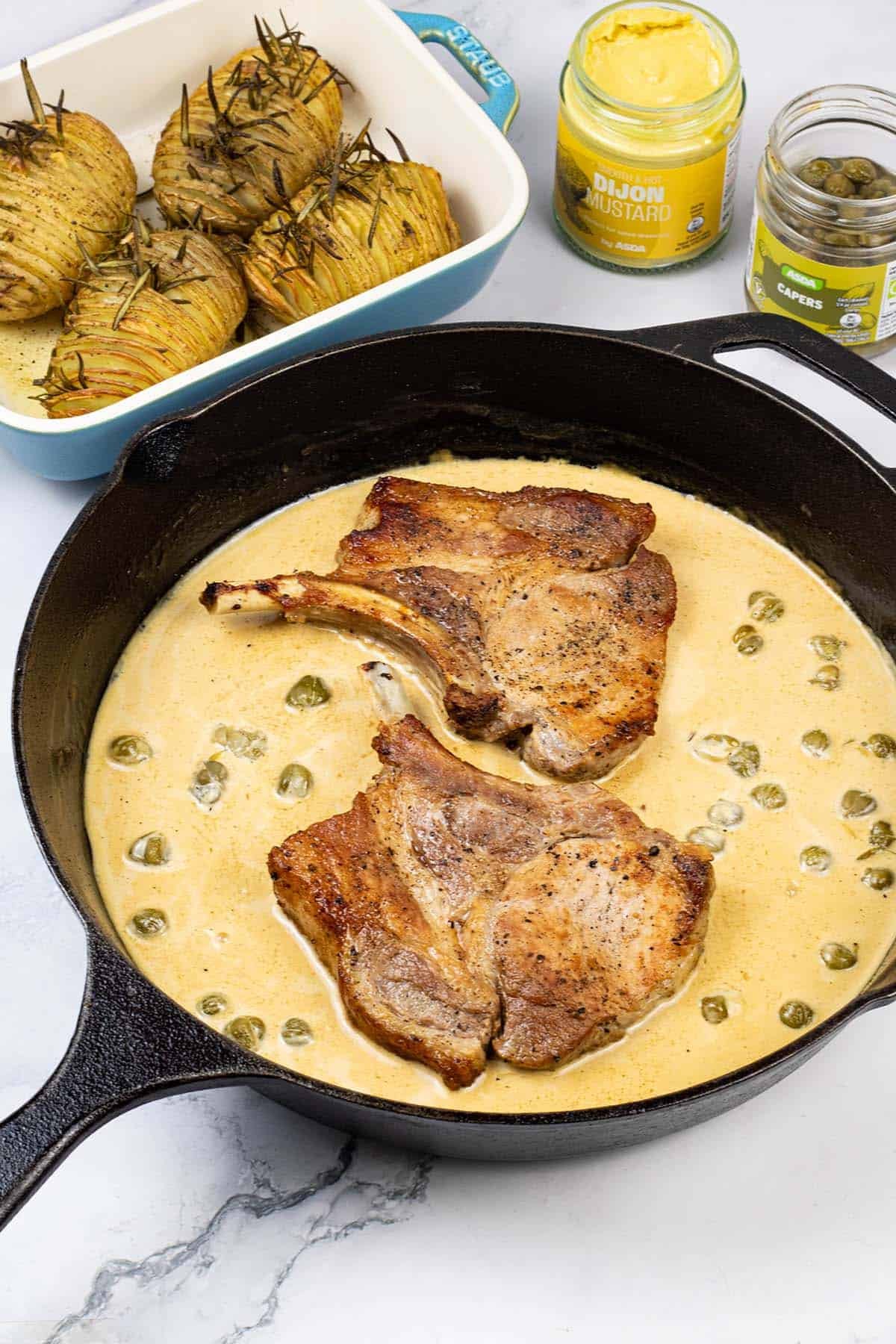 2 portions of pork chops with mustard and capers in a black skillet with garlic and rosemary hasselback potatoes in the background.