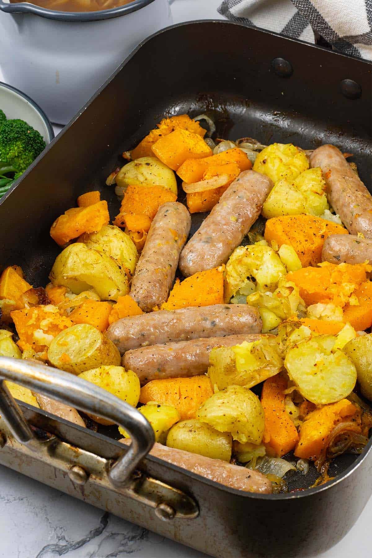 Sausage, squash and potato bake in a large roasting tin with a jug of onion gravy and bowl of broccoli and green beans on the side.