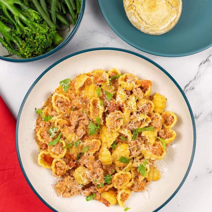 A portion of creamy Italian pork pasta in a teal and cream bowl with a bowl of broccoli and plate of crusty buns on the side.