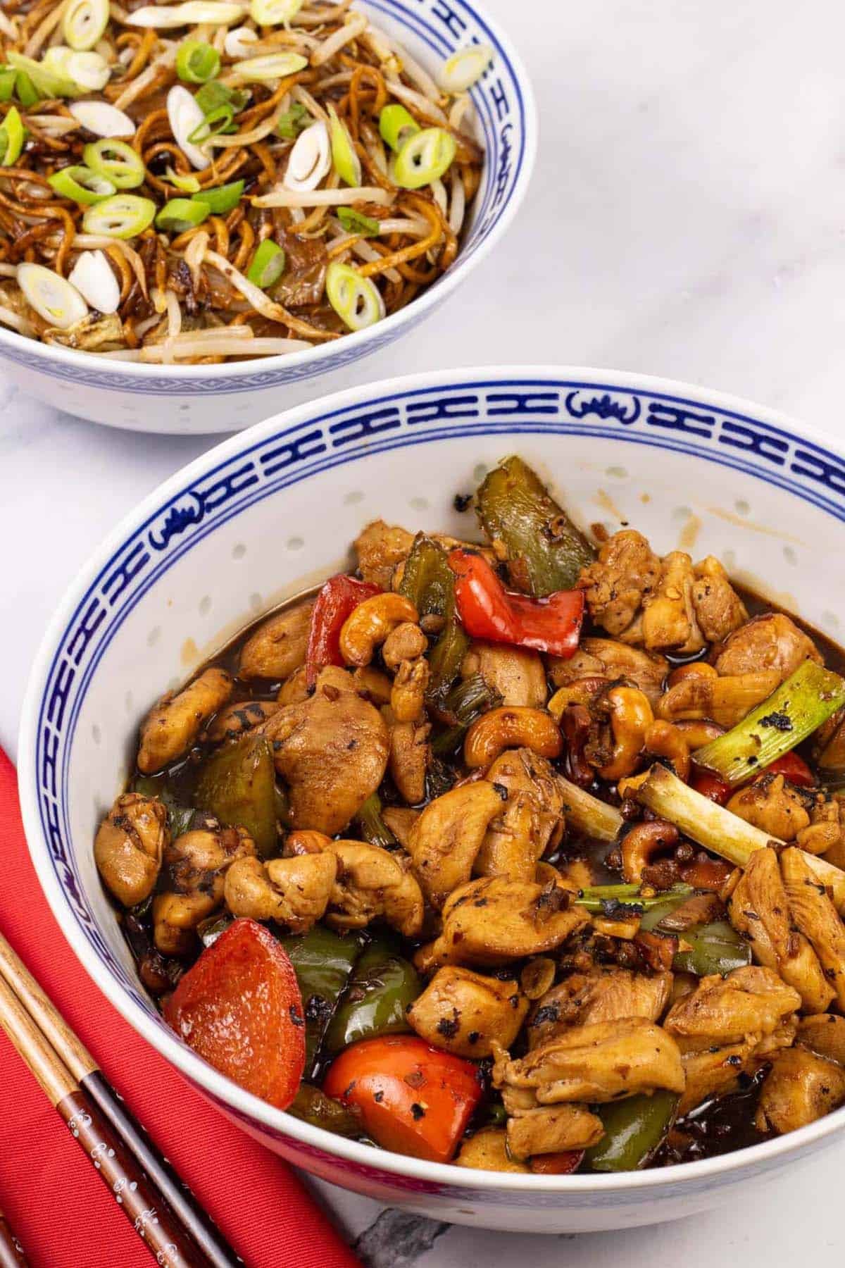 Kung pao chicken in a large blue and white oriental serving bowl with a bowl of fried noodles with beansprouts in the background.