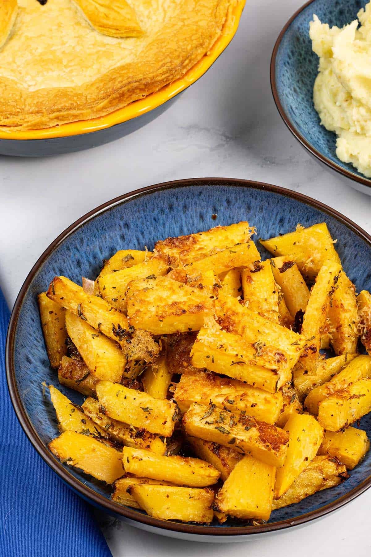 Parmesan and rosemary swede in a blue patterned serving bowl with a pie and mashed potatoes in the background.
