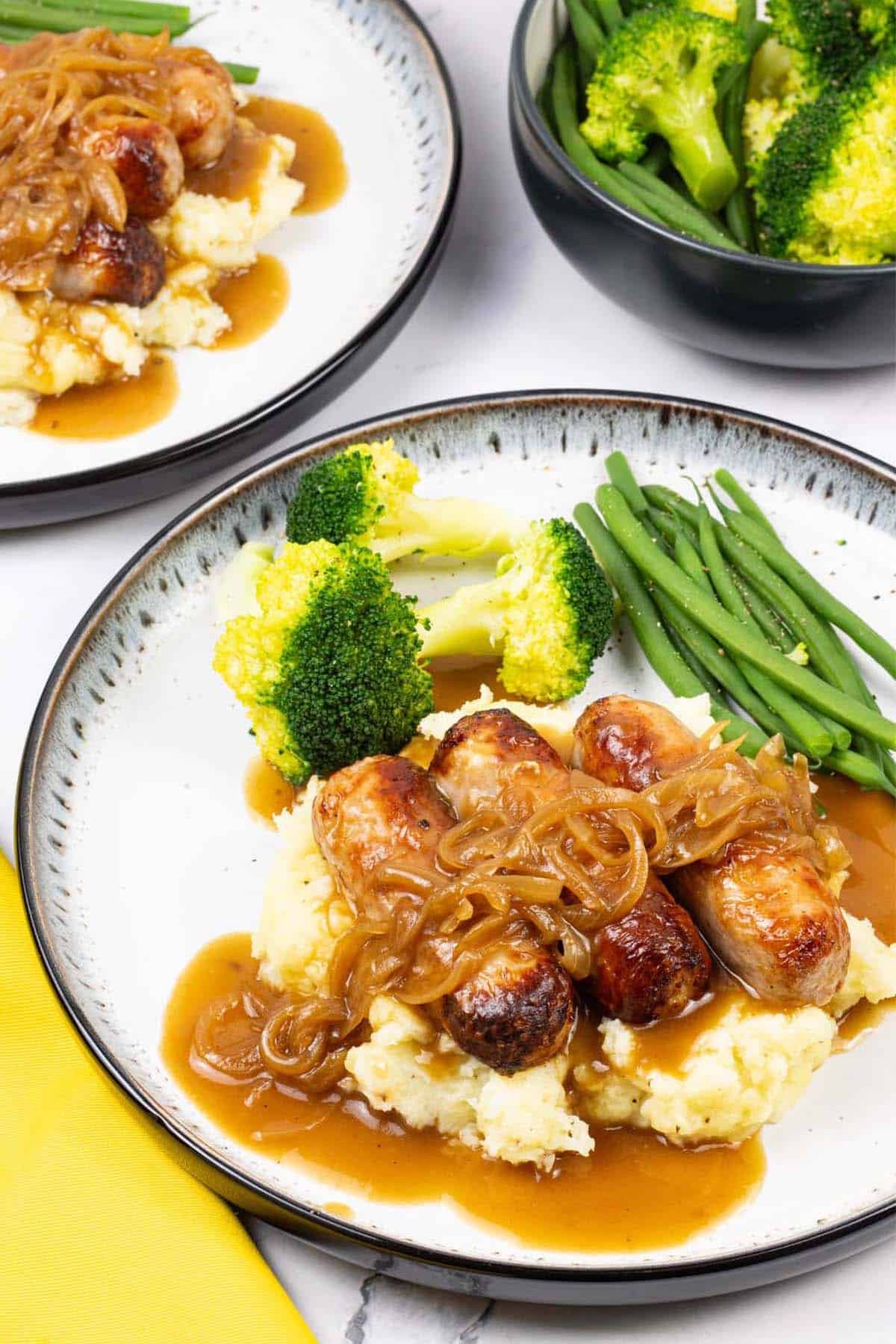 Two plates of sausages with caramelised onion gravy on a bed of mashed potatoes with broccoli and green beans on the side.