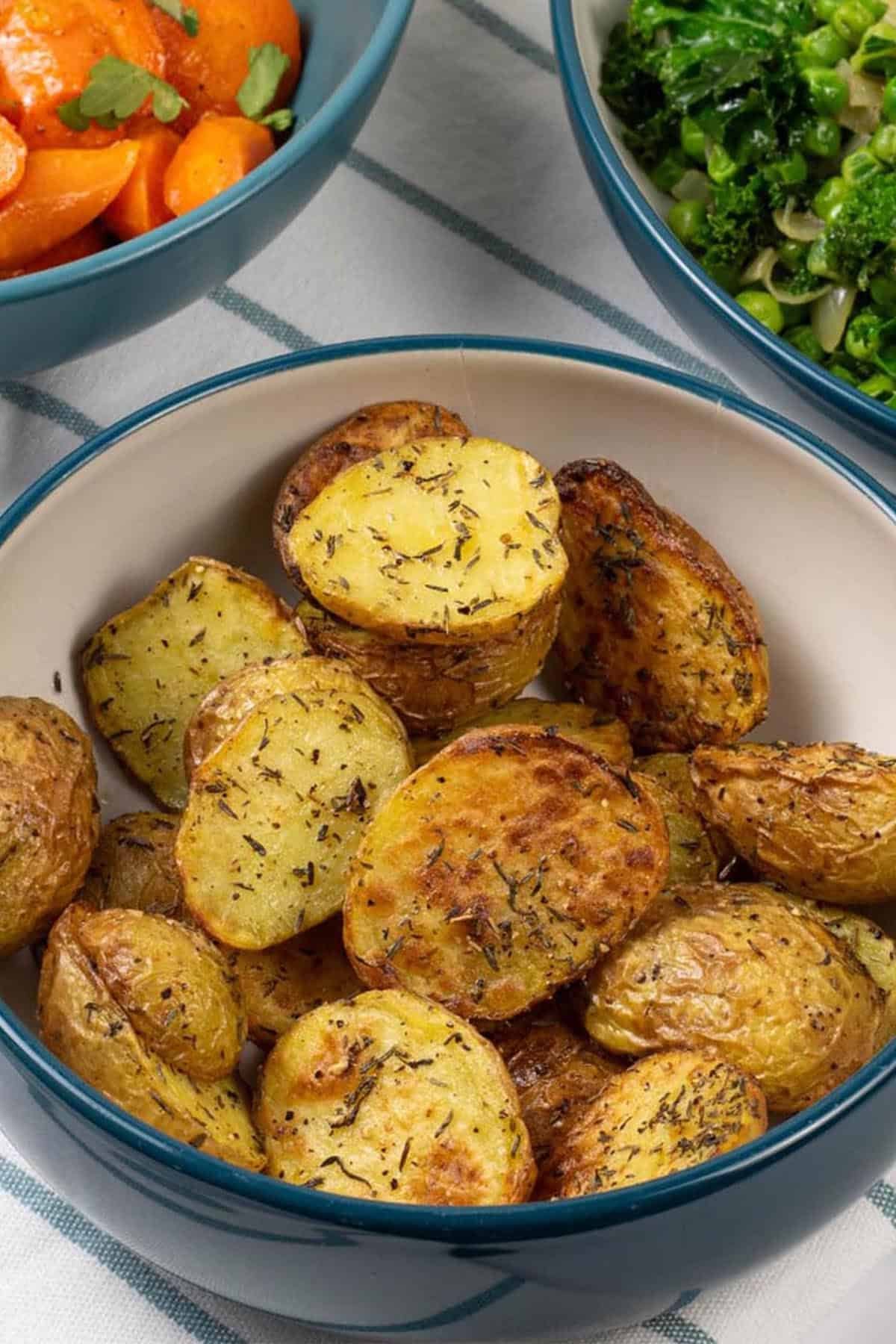 Air fryer thyme roast baby potatoes in a teal and cream serving bowl, with bowls of carrots and kale on the side.
