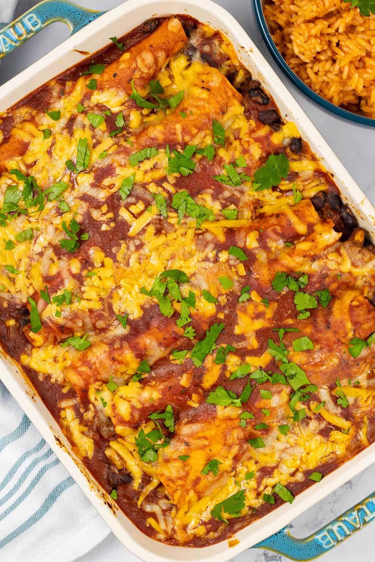 Beef and bean enchiladas in a rectangle Staub ovenproof dish with a bowl of Mexican red rice on the side.