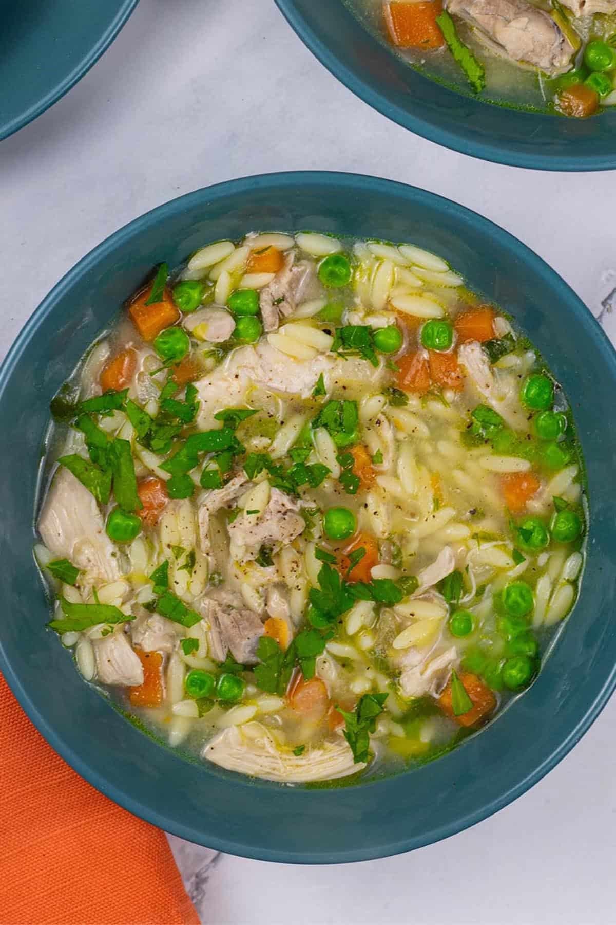A portion of chicken orzo soup in a teal bowl with an orange napkin on the side.
