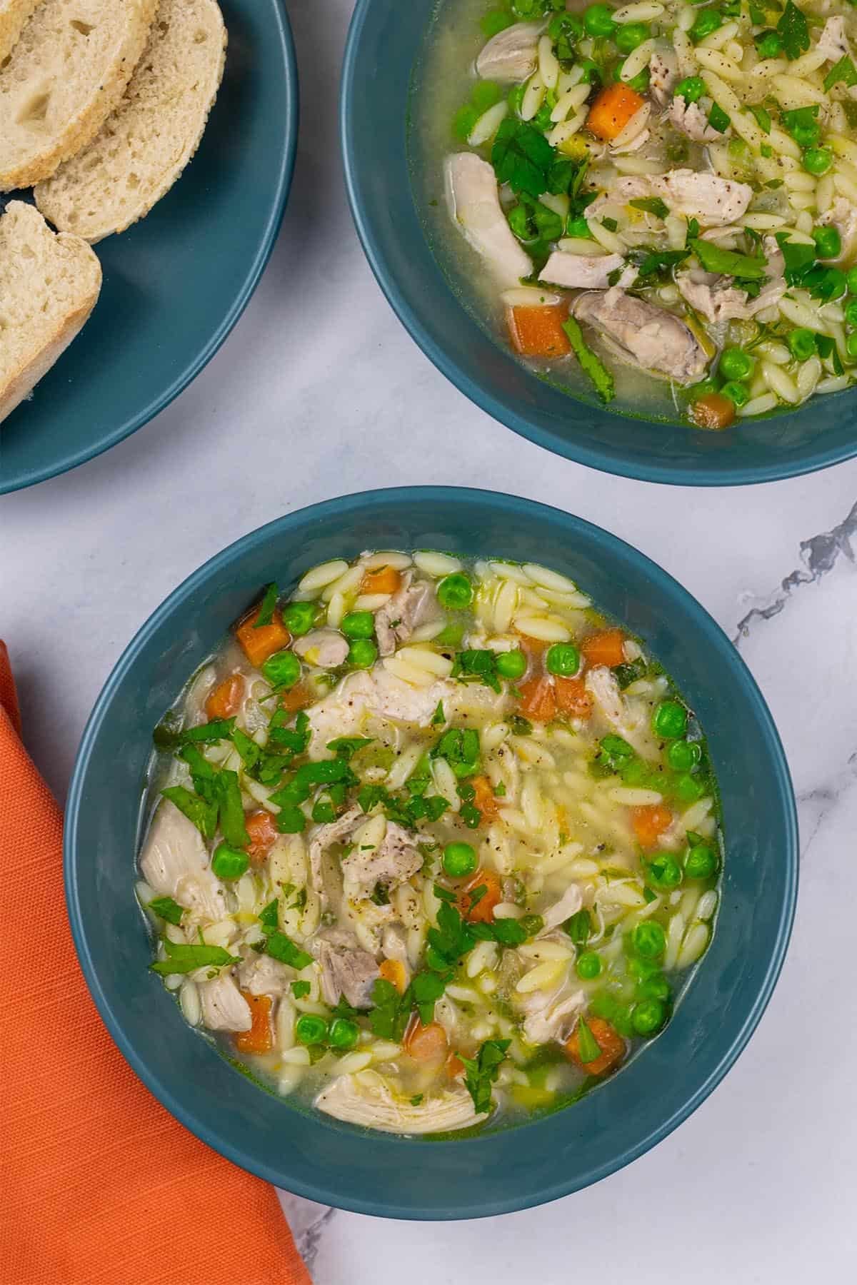 Two portions of chicken orzo soup in teal bowls with a plate of sliced crusty bread on the side.