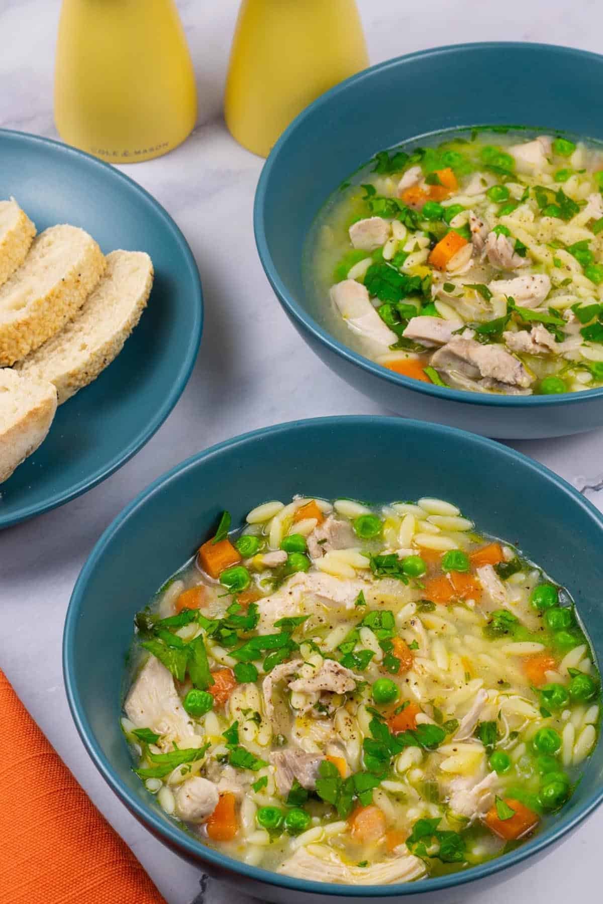 Two portions of chicken orzo soup in teal bowls with a plate of sliced crusty bread on the side.