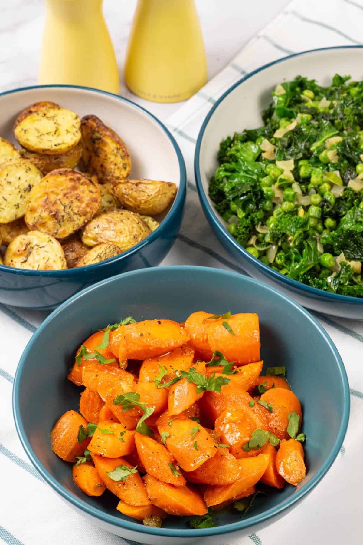 Marmalade roast carrots in a teal serving bowl with bowls of air fryer roast baby potatoes and kale on the side.