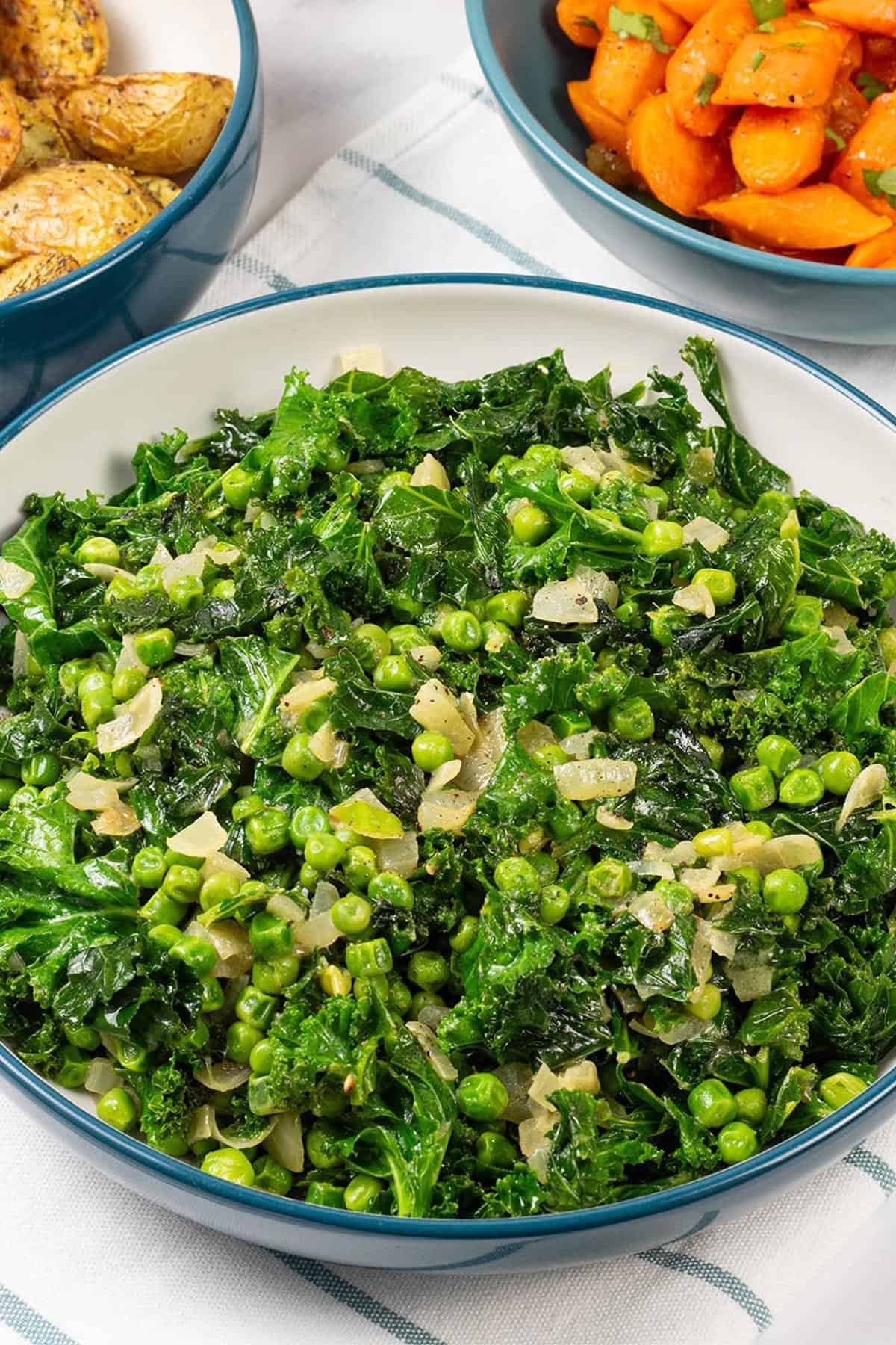 Petit pois with kale and onions in a large teal and cream serving bowl, with bowls of carrots and air fryer roast baby potatoes on the side.