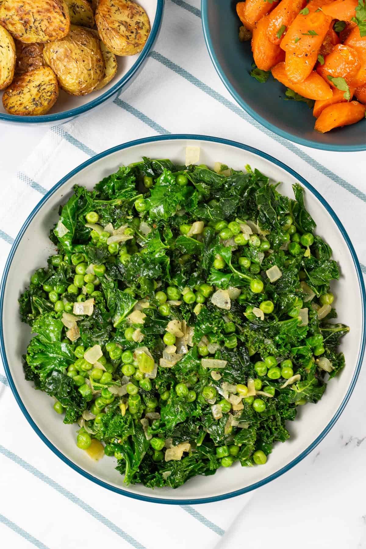 Petit pois with kale and onions in a large teal and cream serving bowl, with bowls of carrots and air fryer roast baby potatoes on the side.