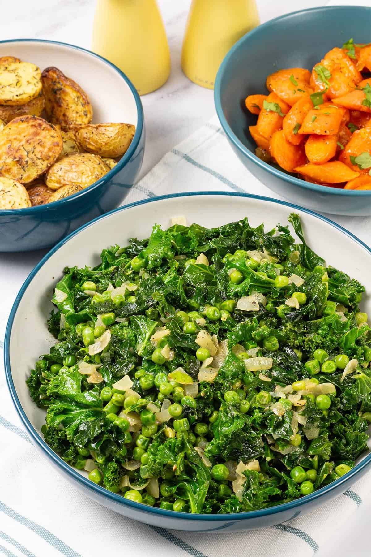 Petit pois with kale and onions in a large teal and cream serving bowl, with bowls of carrots and air fryer roast baby potatoes on the side.