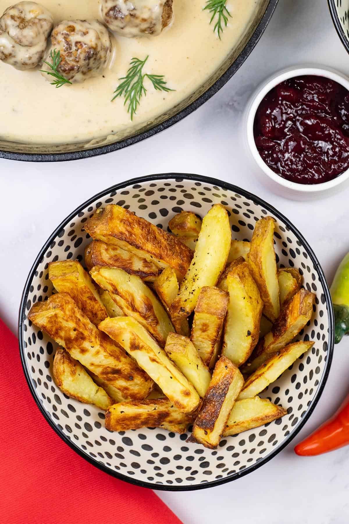 Chunky oven chips in a small serving bowl with scandi-style meatballs with gravy and a small dish of lingonberry spread on the side.