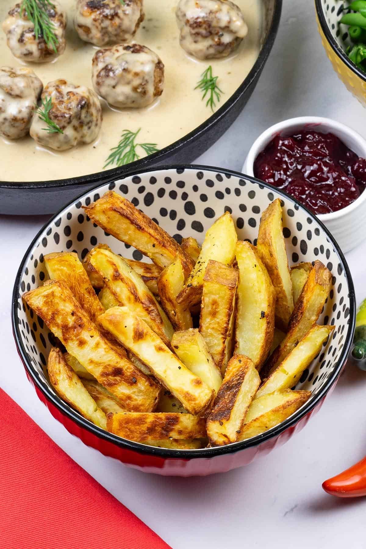 Chunky oven chips in a small serving bowl with scandi-style meatballs with gravy and a small dish of lingonberry spread on the side.