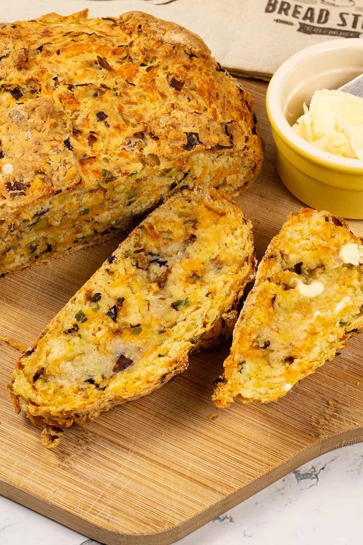 Leek and red leicester soda bread on a board with a couple of slices cut, and a small dish of butter and bread bag on the side.