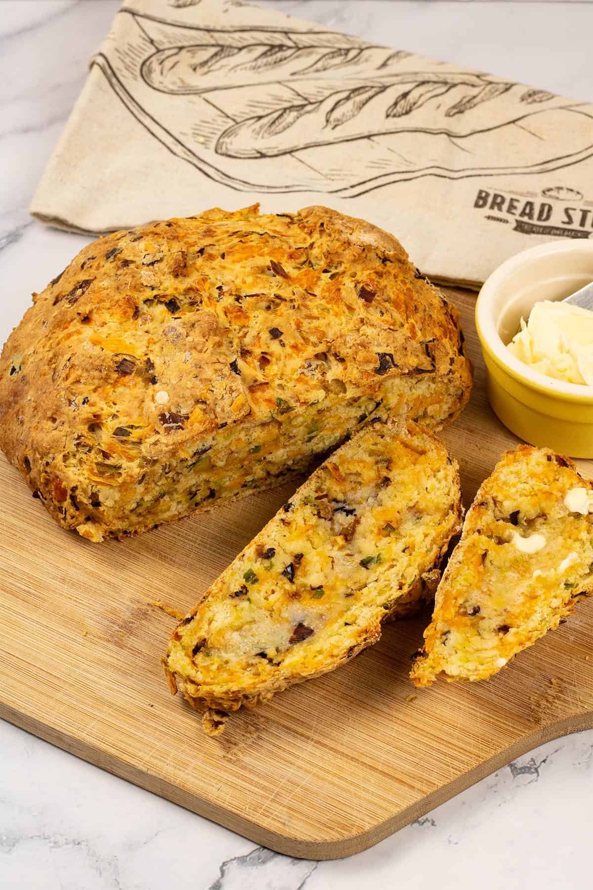 Leek and red leicester soda bread on a board with a couple of slices cut, and a small dish of butter and bread bag on the side.