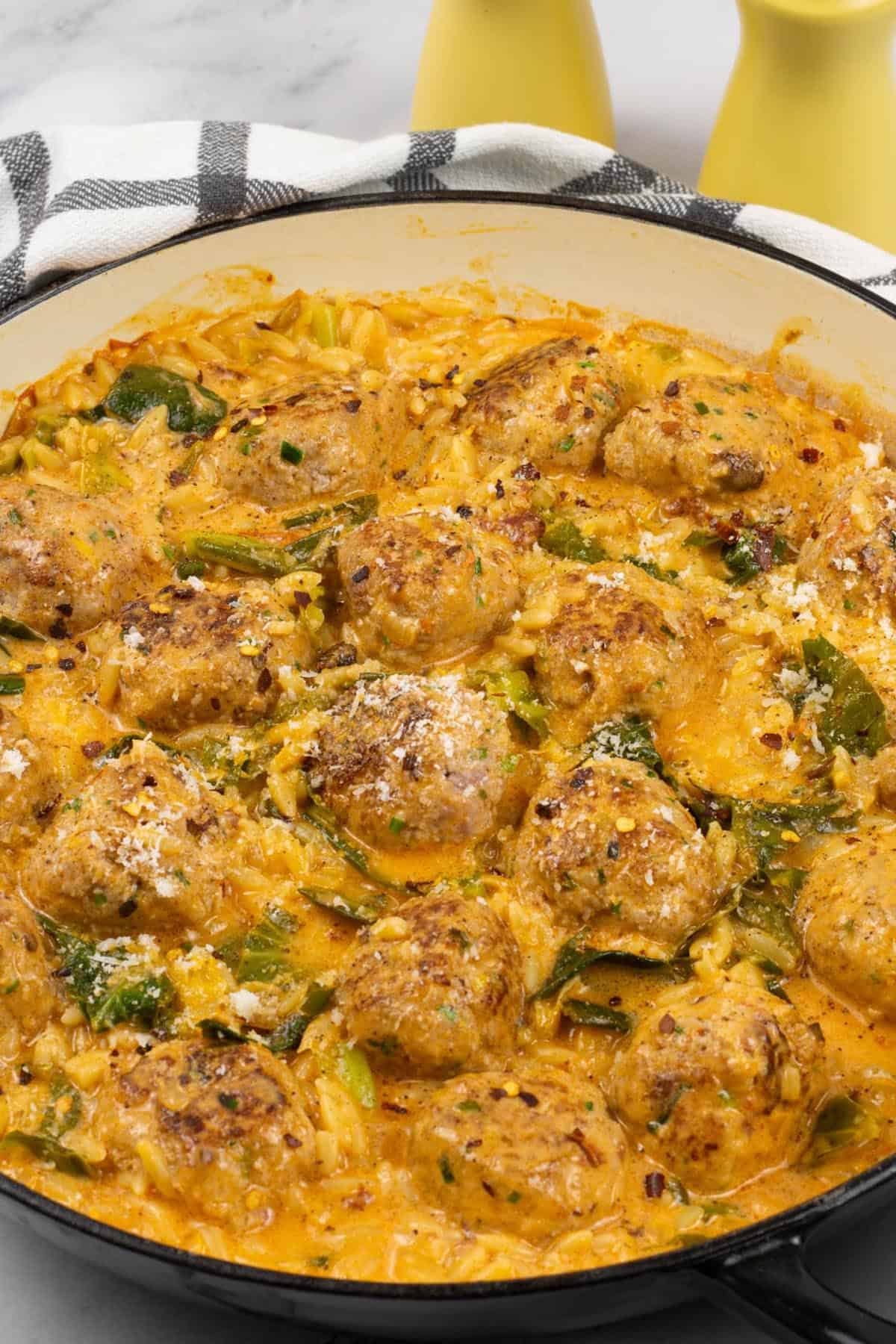 Marry me chicken meatball orzo in a large shallow casserole dish, with a black and white checked towel and yellow salt and pepper grinders on the side.