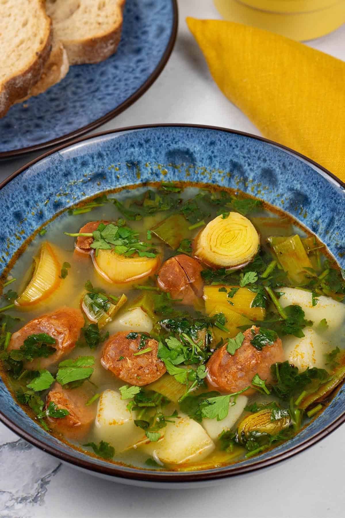 A portion of potato, leek and chorizo soup in a blue patterned bowl with a plate of bread, small dish of butter and mustard napkin on the side.