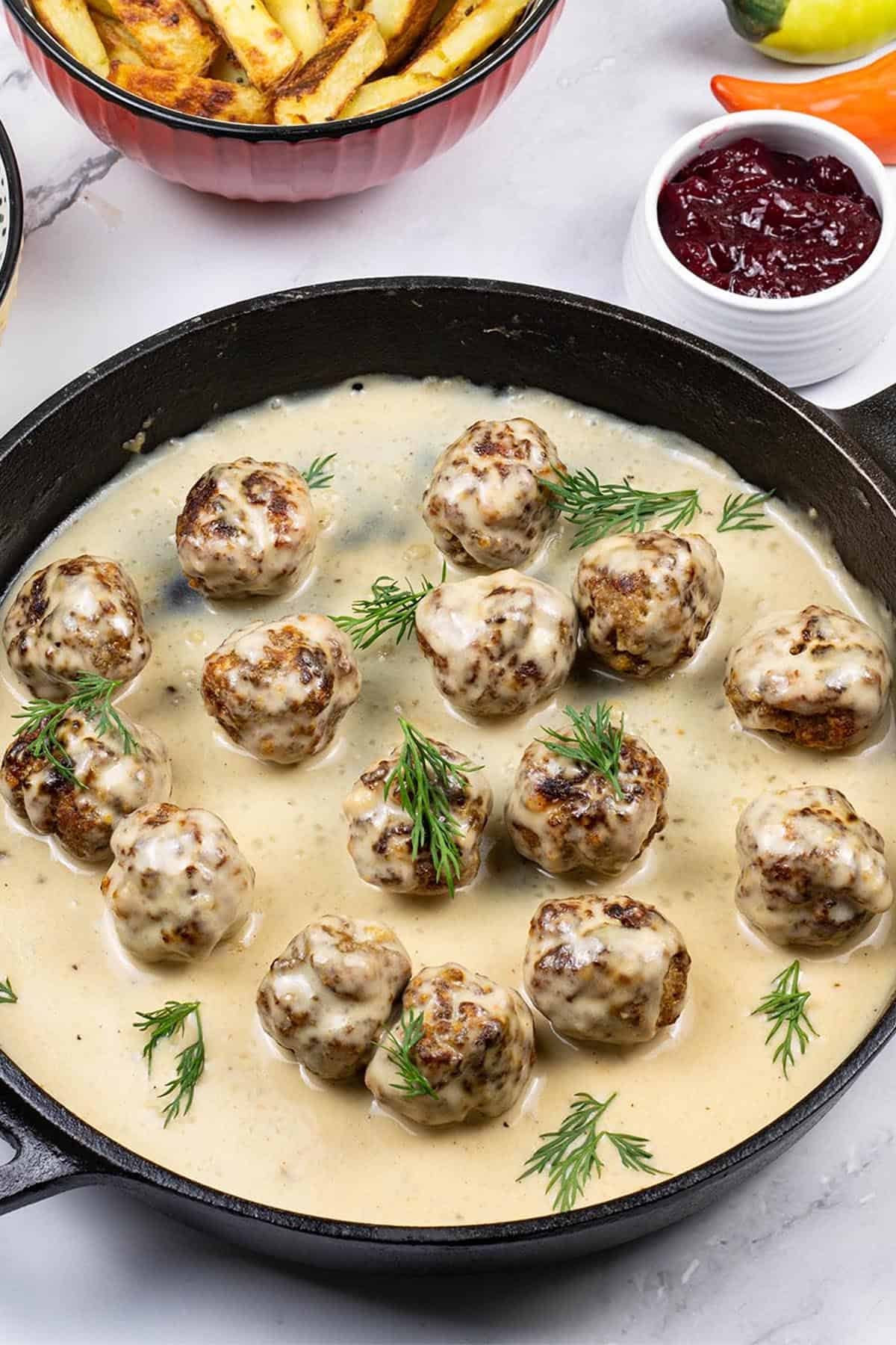 Scandi-style meatballs with gravy in a skillet, with a small dish of lingonberry spread on the side.