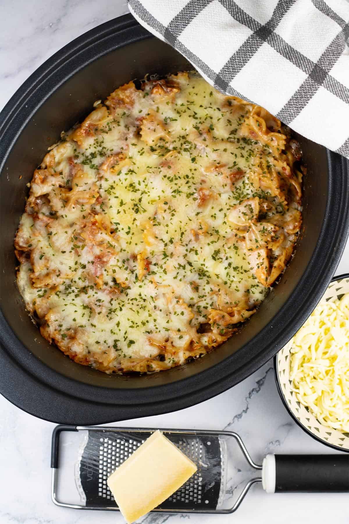 Slow cooker chicken parmesan pasta in the slow cooker with a block of parmesan and grater, and bowl of mozzarella on the side.