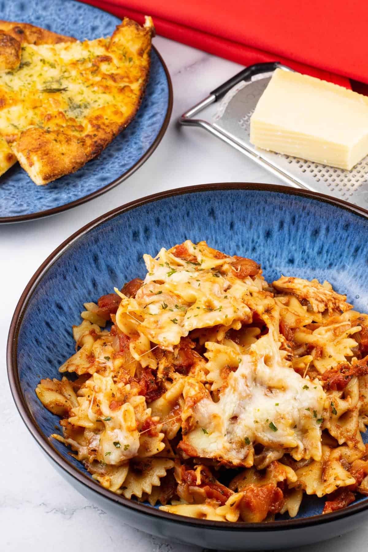 A portion of slow cooker chicken parmesan pasta in a blue patterned bowl with a plate of garlic bread, block of parmesan cheese and a red napkin on the side.