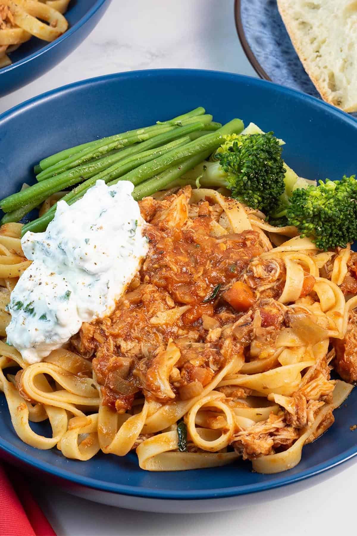 Close up of a portion of slow cooker chicken ragu with basil ricotta in a blue bowl, with green beans and broccoli on the side.