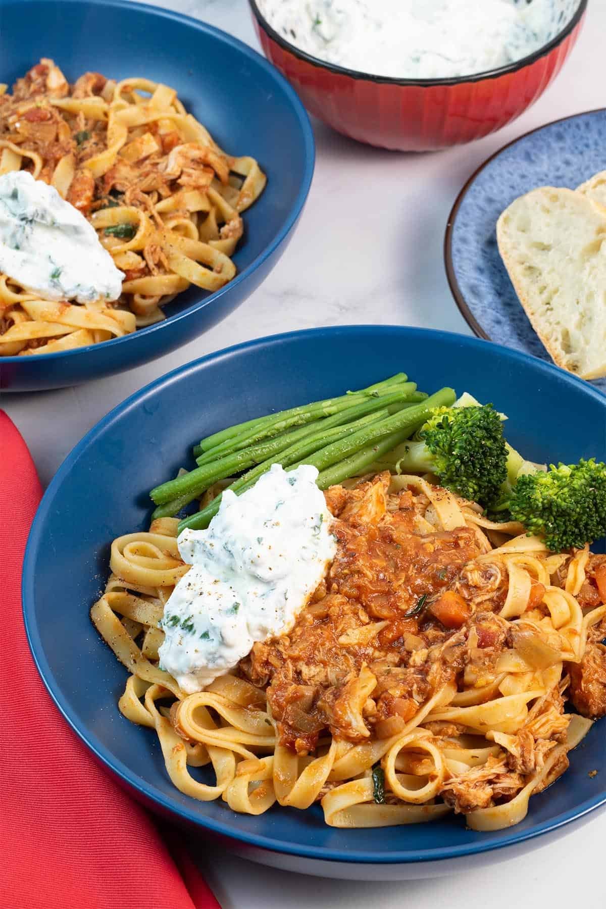 Two portions of slow cooker chicken ragu with basil ricotta in blue bowls, with a bowl of basil ricotta and plate of sliced crusty bread on the side.
