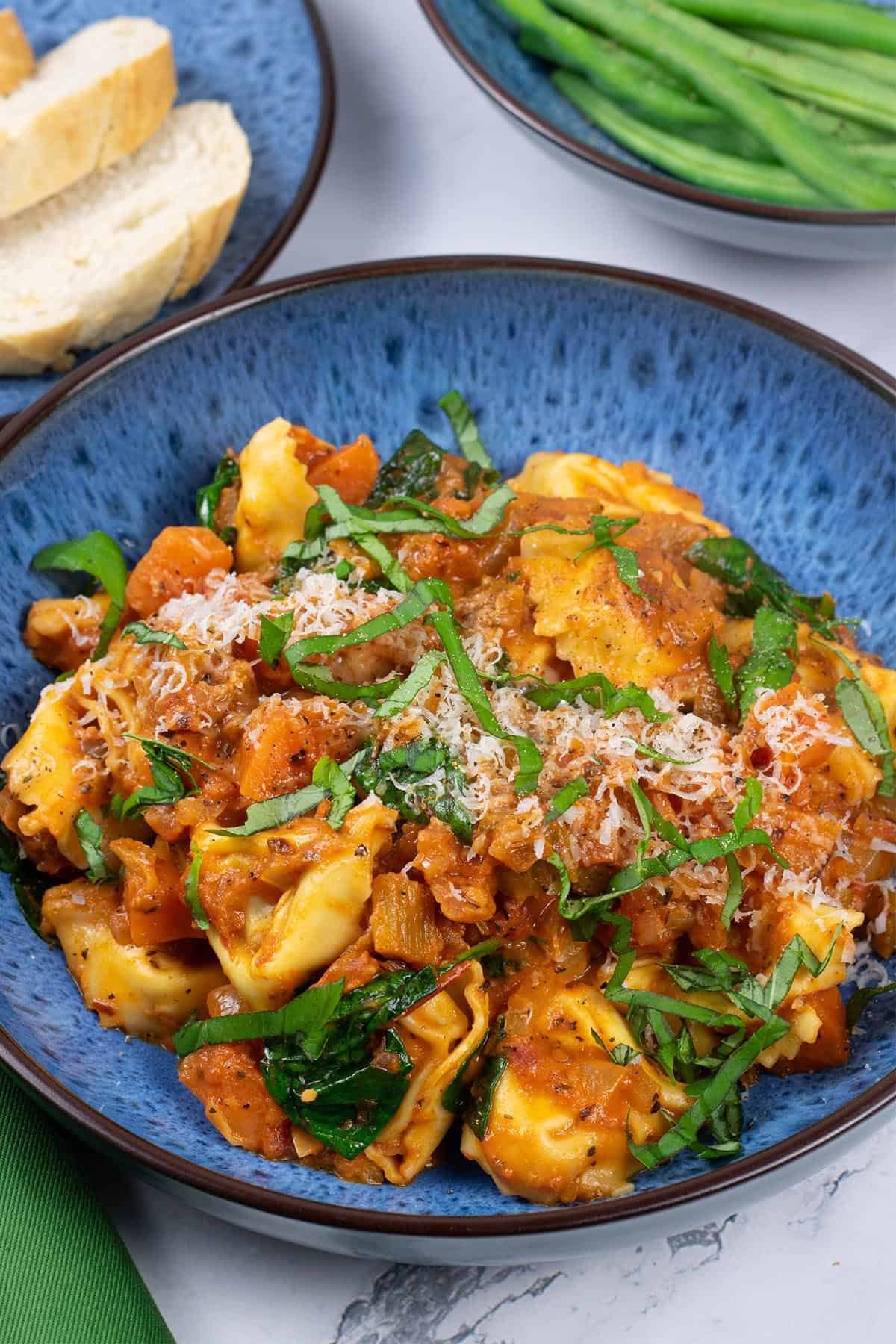 A portion of tortellini with creamy tomato and spinach sauce in a blue patterned bowl with a bowl of green beans and plate of sliced bread on the side.